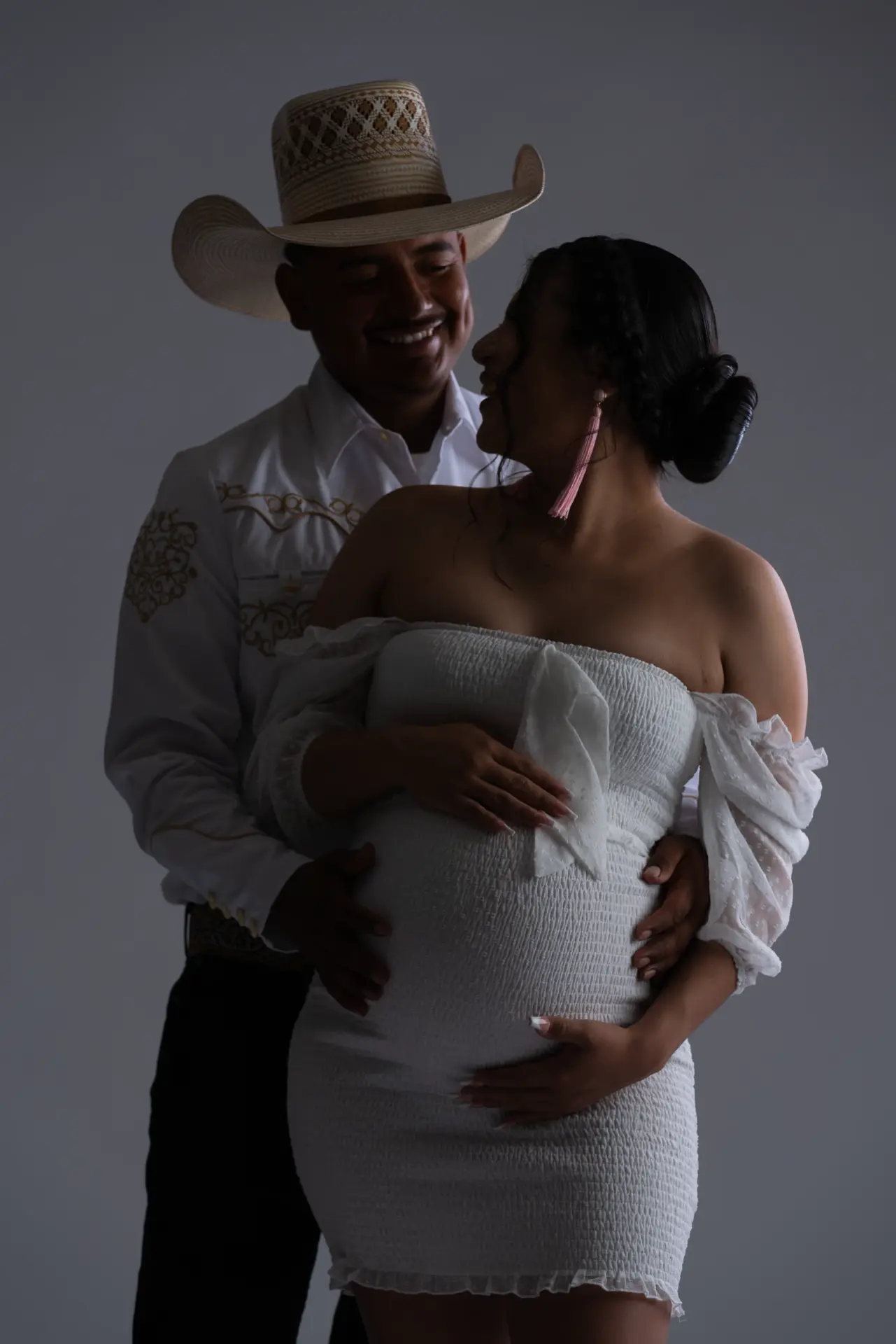Expecting couple in white with western hats, moody studio maternity portrait