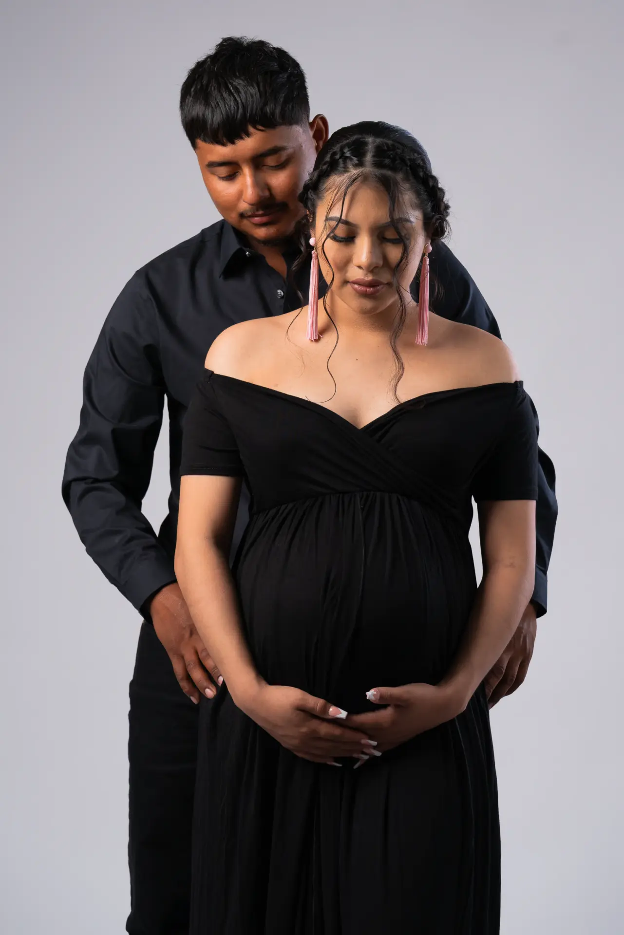 Expecting couple in black embracing in a gray studio, his hands on her bump