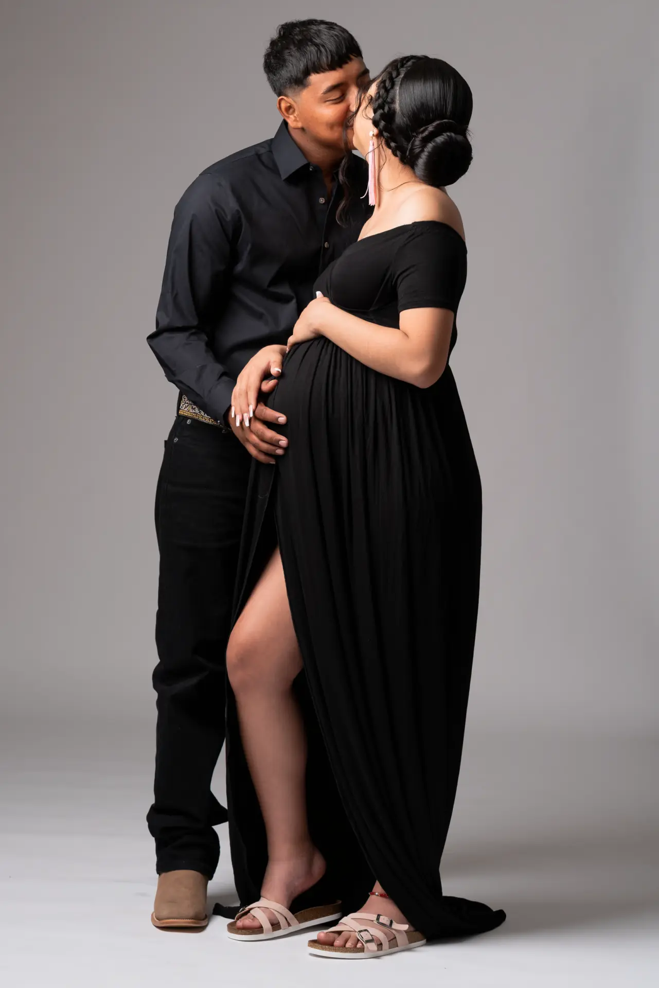 Expecting couple standing together in black, partner's arm around her bump