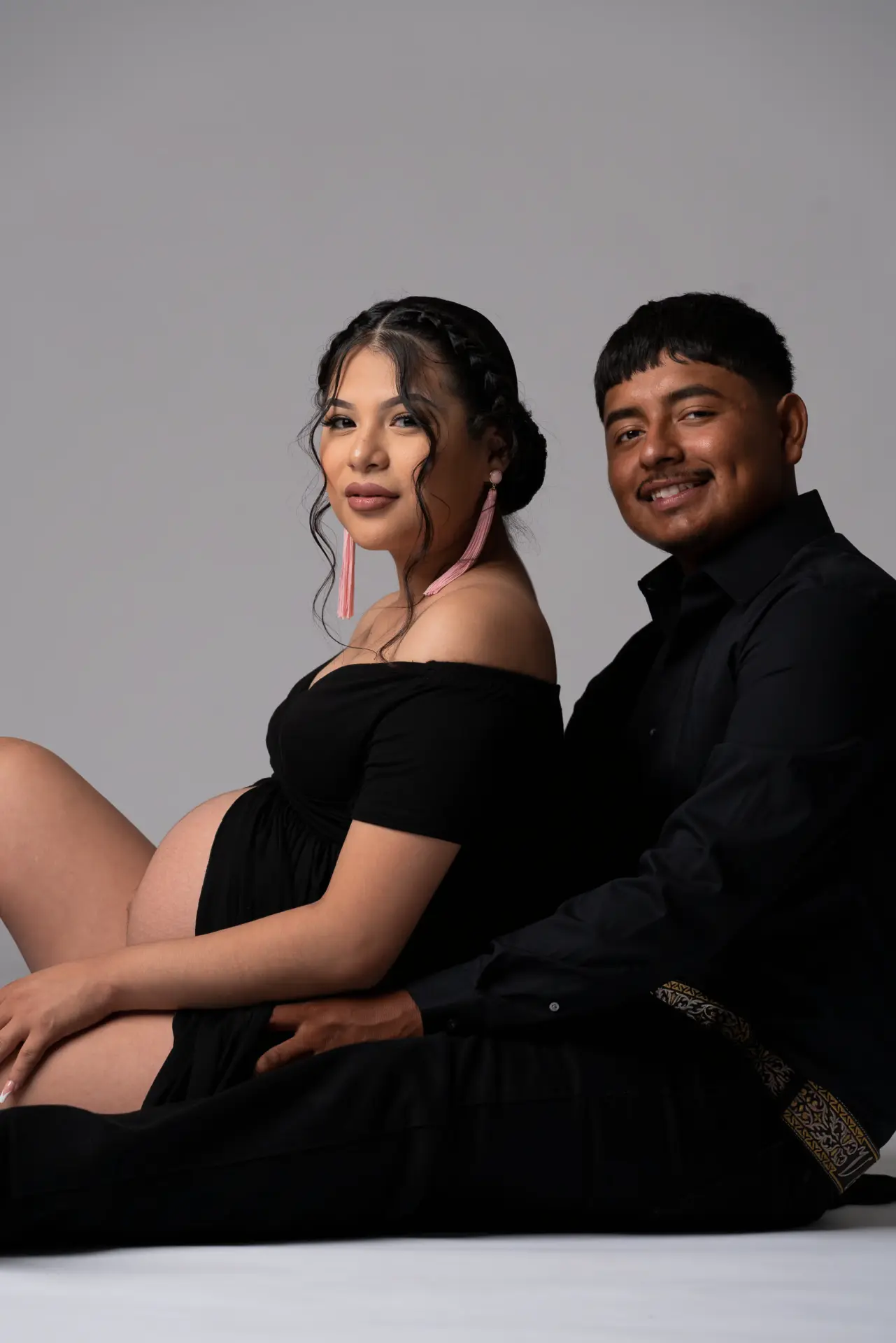 Expecting couple seated together on a gray studio floor in black outfits