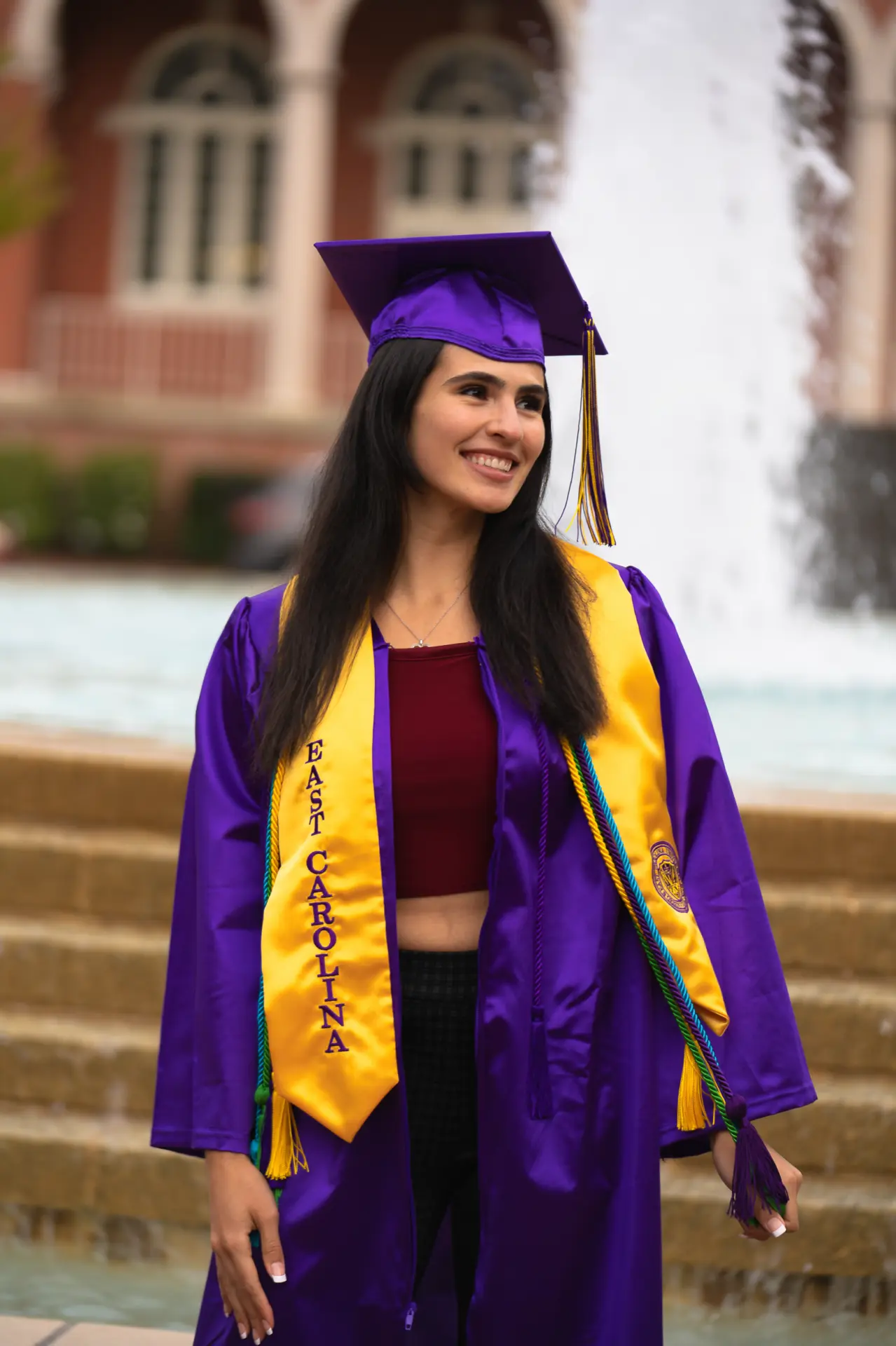Graduate in a purple gown, cap and gold stole in front of a brick hall