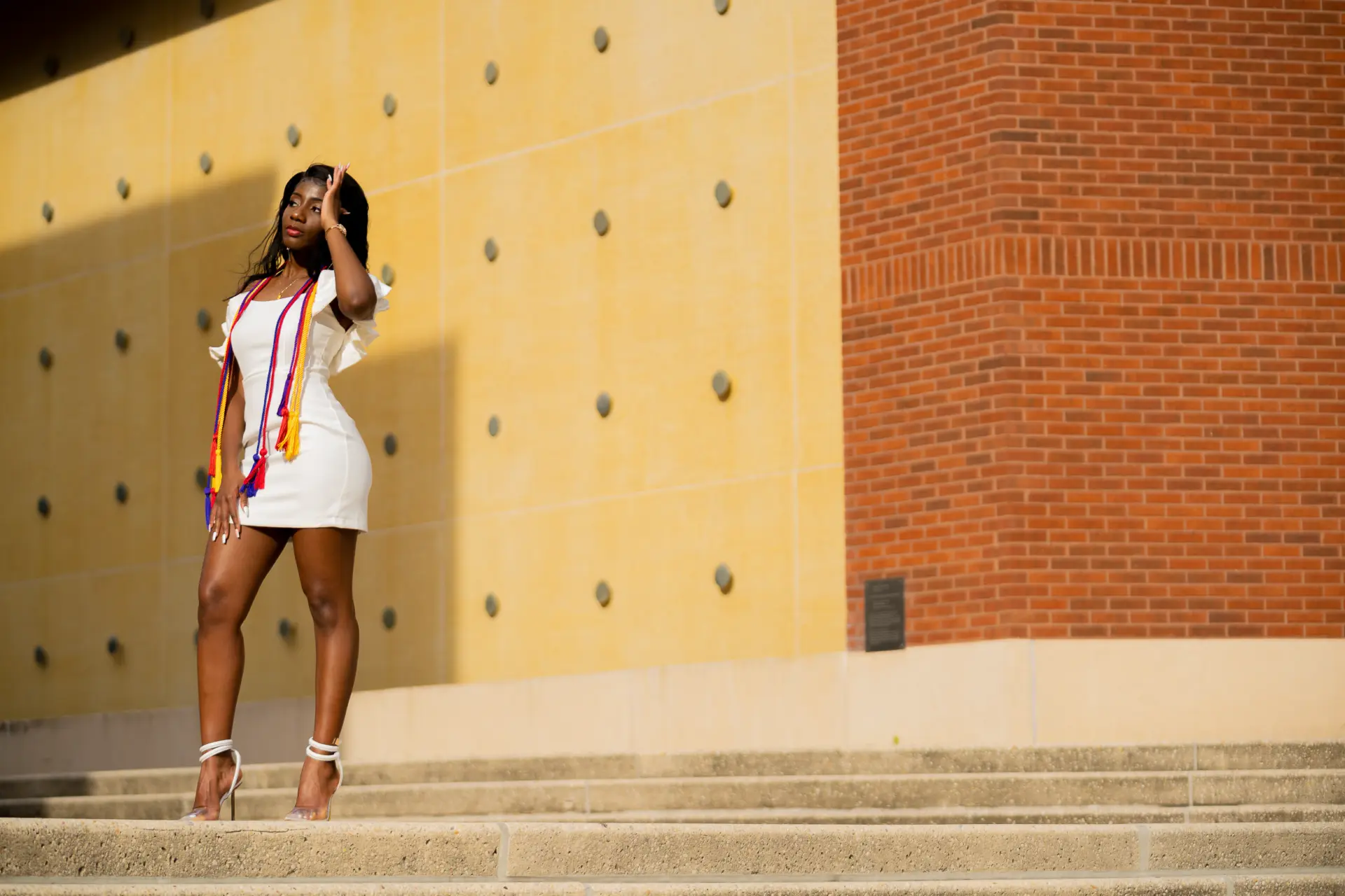 Graduate in white posing against a sunlit campus wall