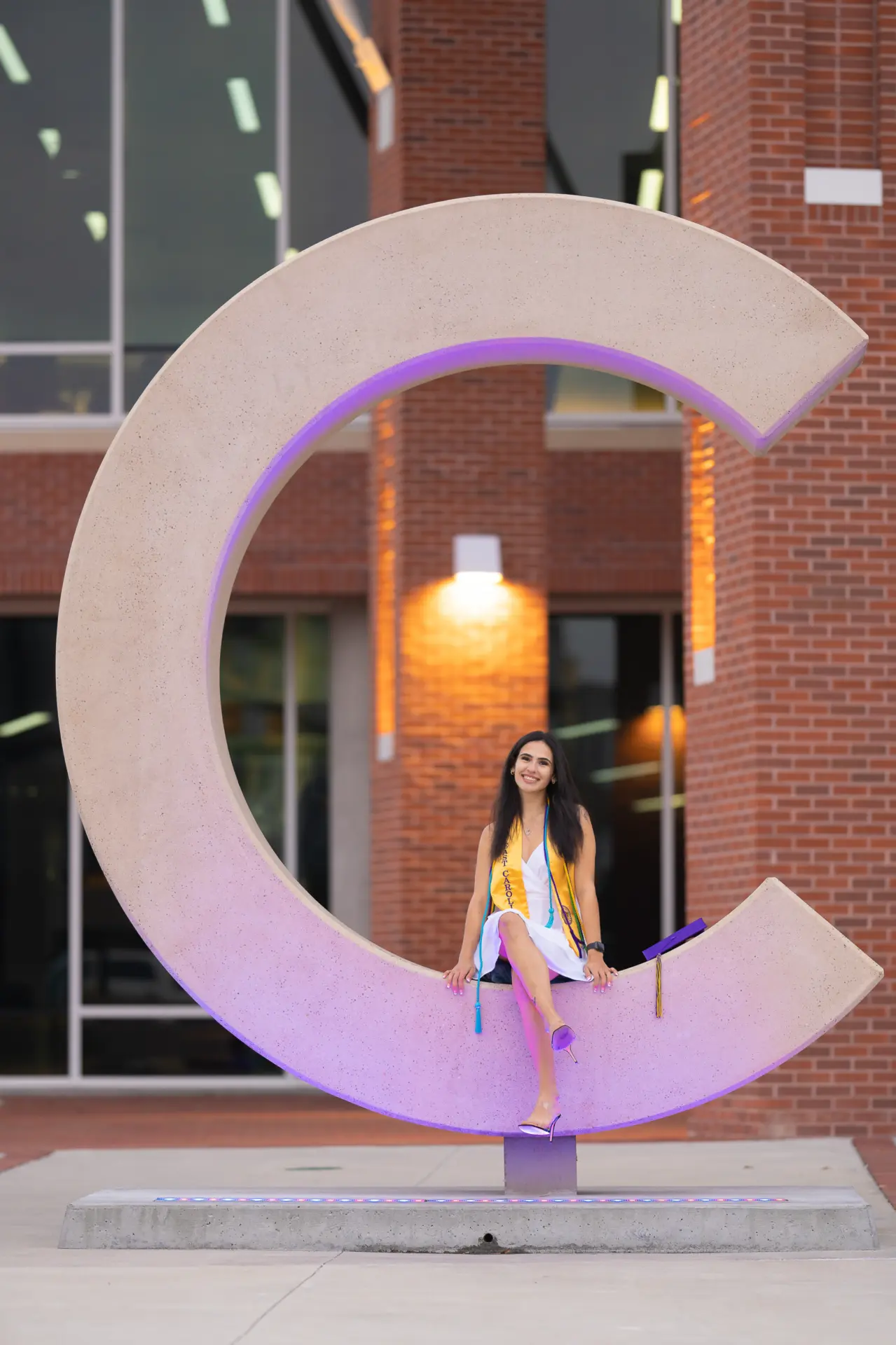 Graduate posing within the ECU C sculpture lit purple at dusk
