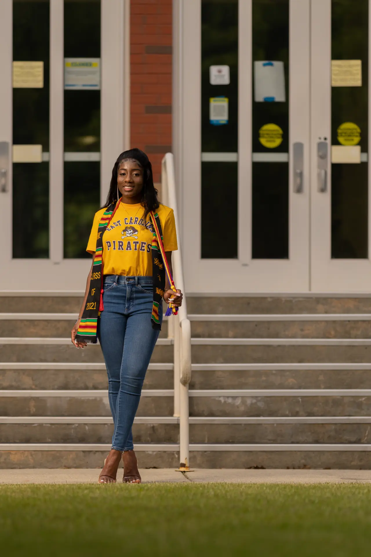 Graduate in a gold shirt and jeans on the steps of a campus building