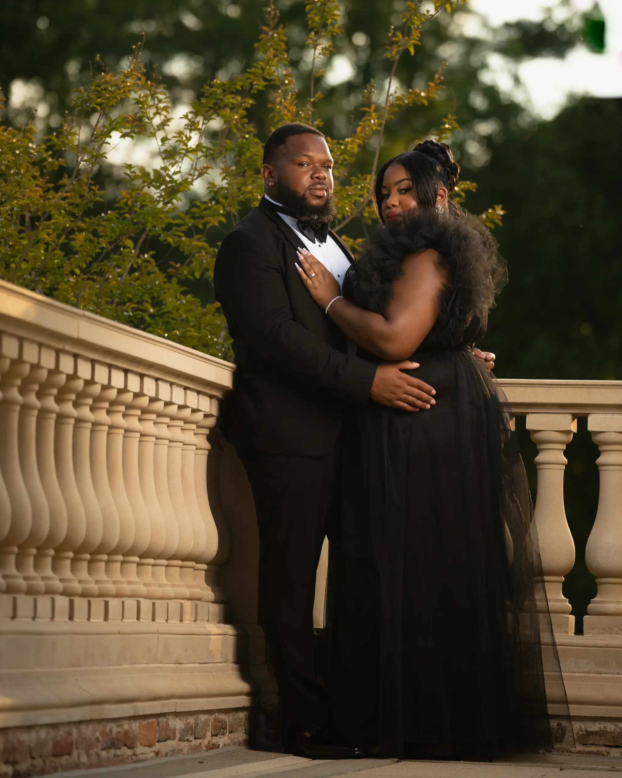 Couple in black formal wear embraces on a stone balcony with trees behind