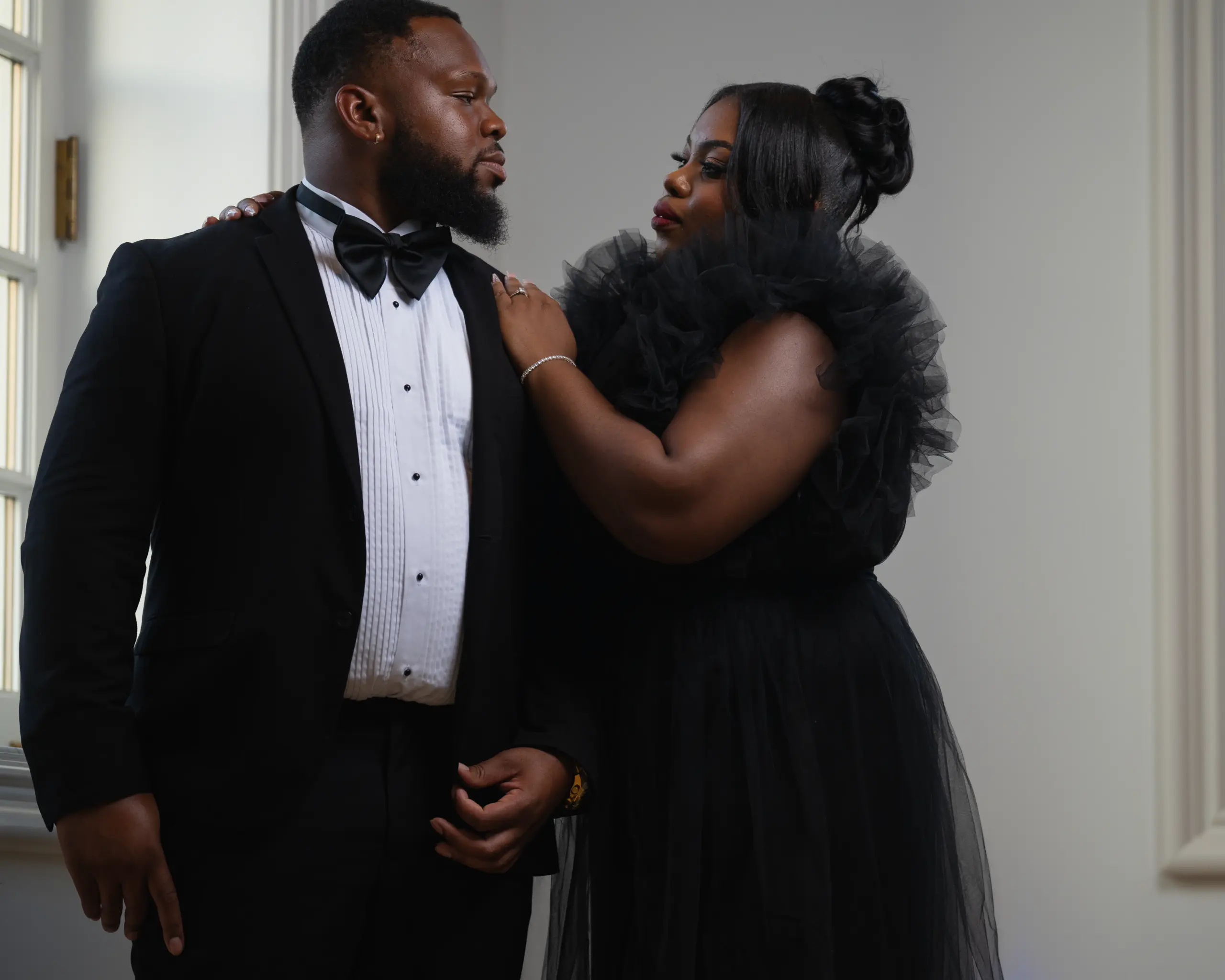 Couple in black formal wear poses by a large window in soft light