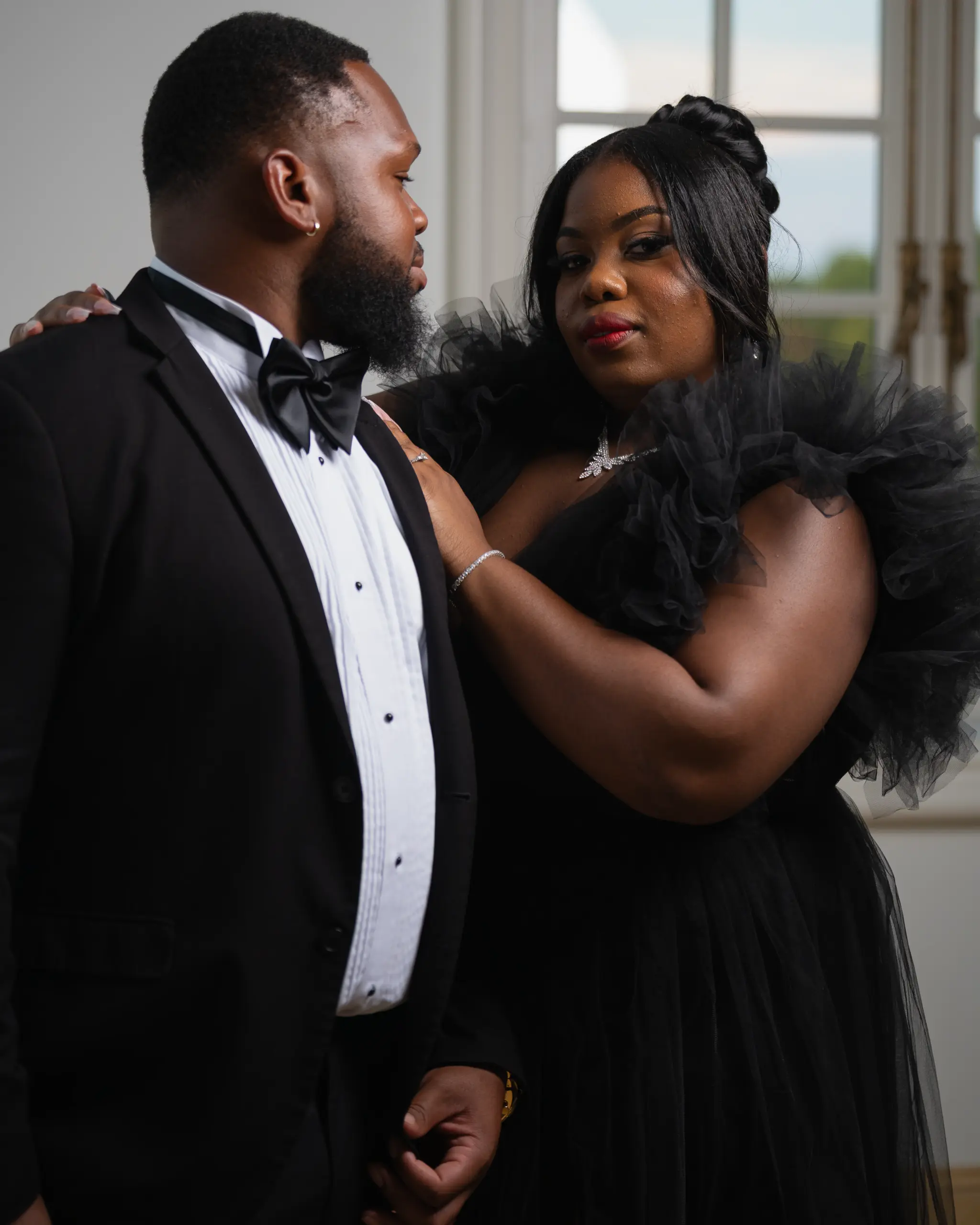 Man in a tuxedo holds his partner in a black gown by a window