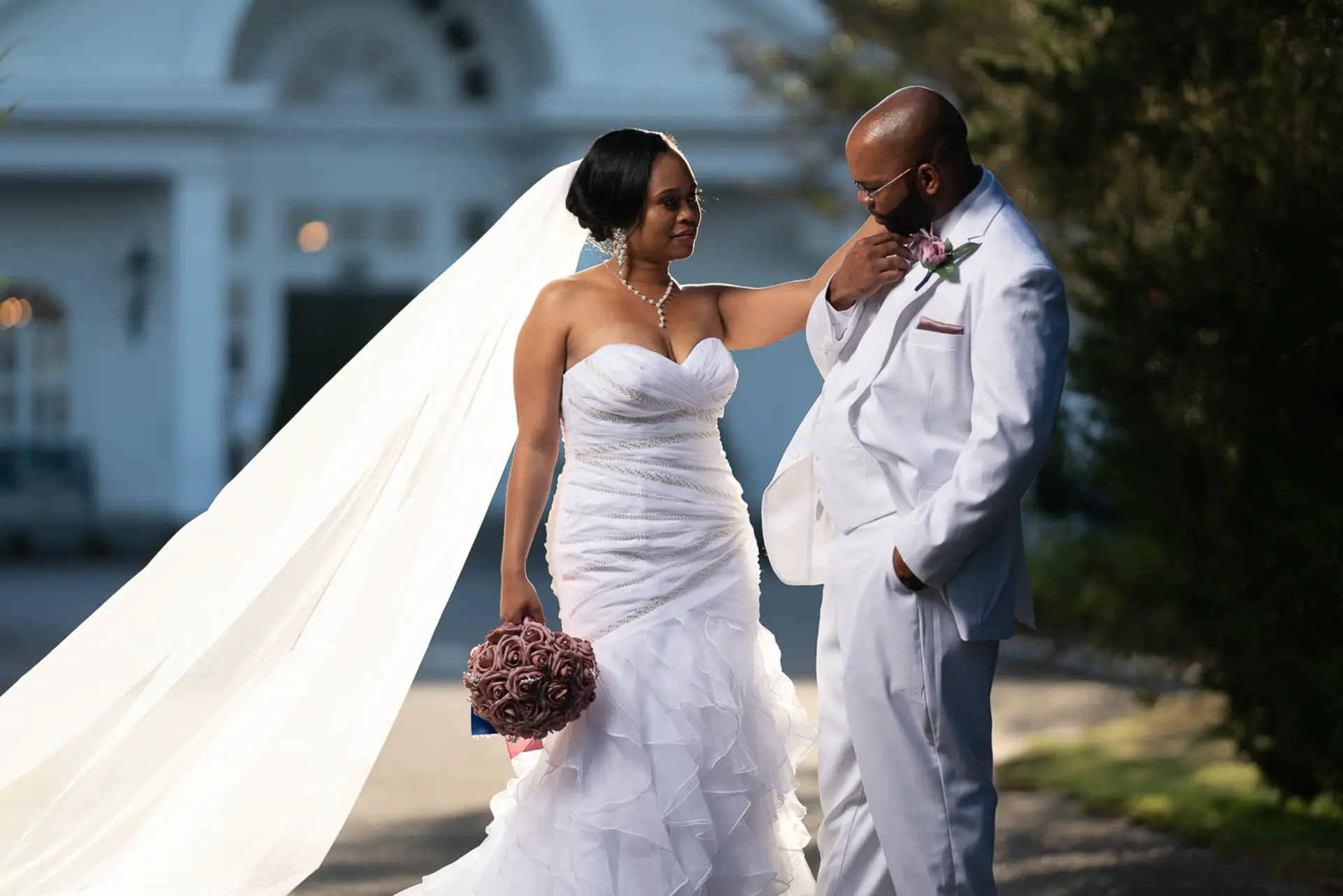 Bride and groom posing in front of a white columned estate with her veil flowing