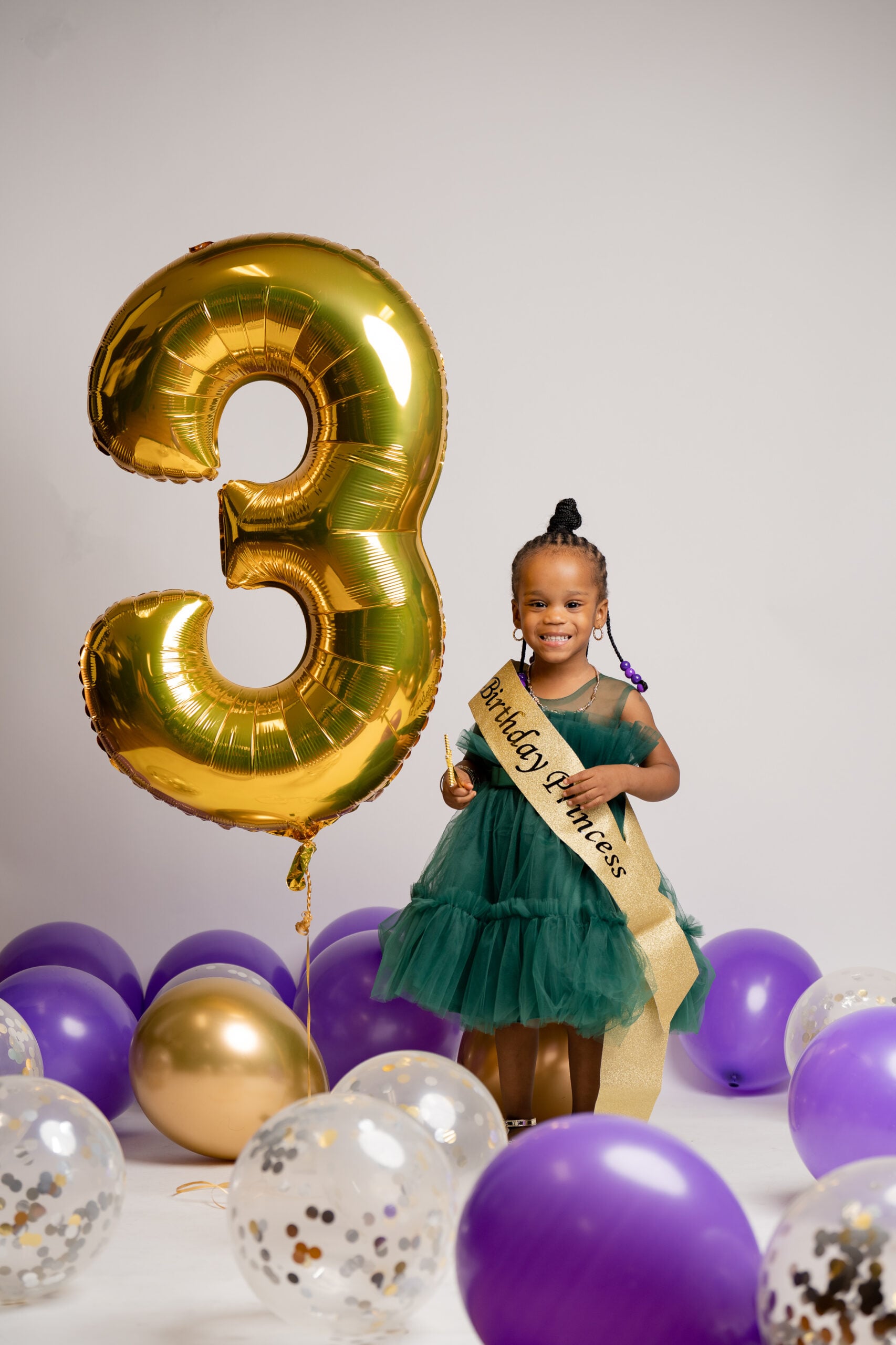 Little girl in a green tutu with gold "3" balloon and purple balloons