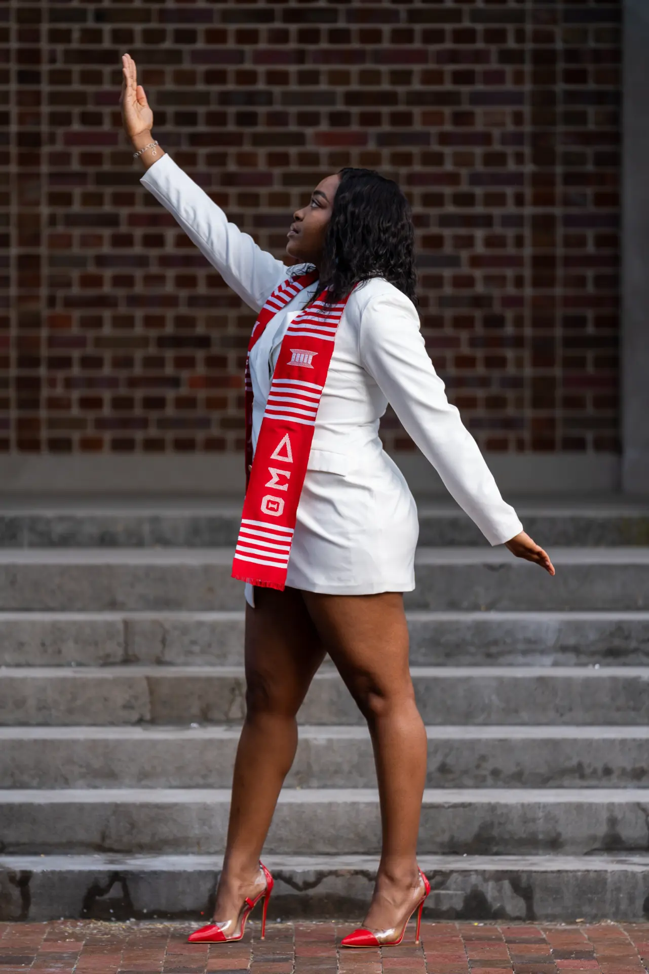 Graduate in white with a red stole raising her arm on brick steps