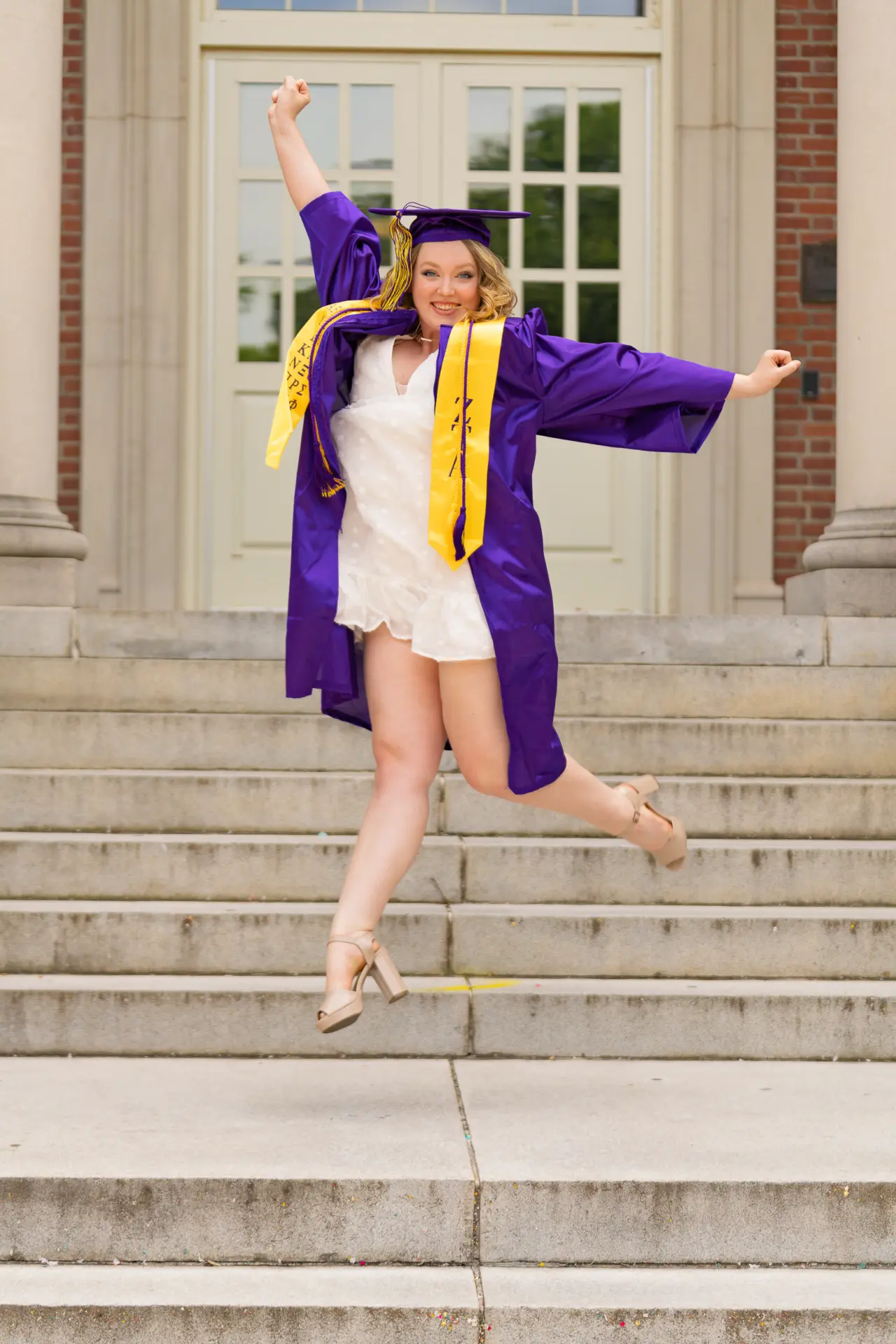 Graduate in a purple gown celebrating on the steps of a white campus building