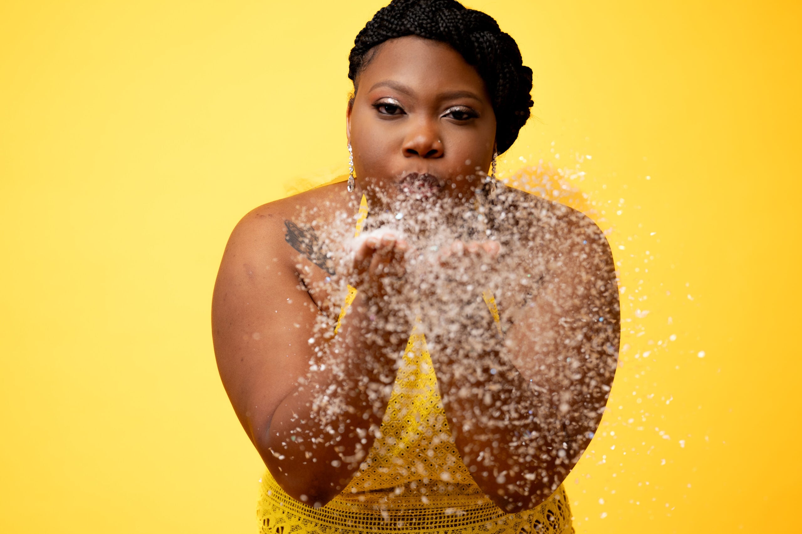 Woman blowing gold glitter toward the camera on a yellow backdrop