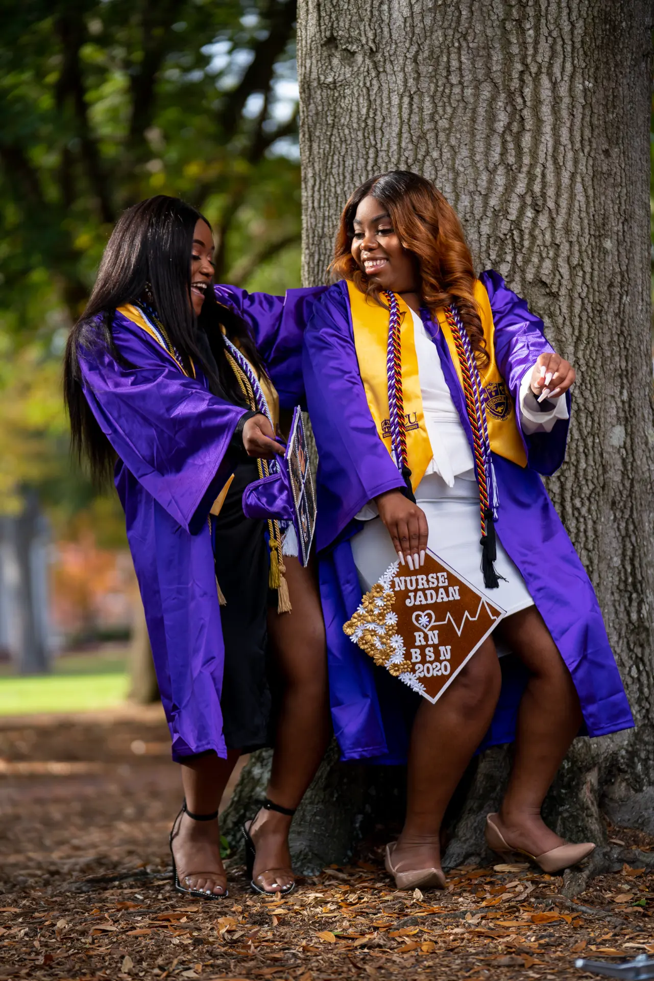Two graduates in purple gowns laughing together under a large tree
