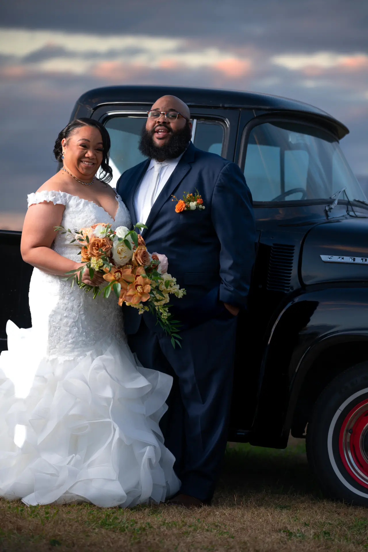 Groom holds the bride beside a classic truck at sunset