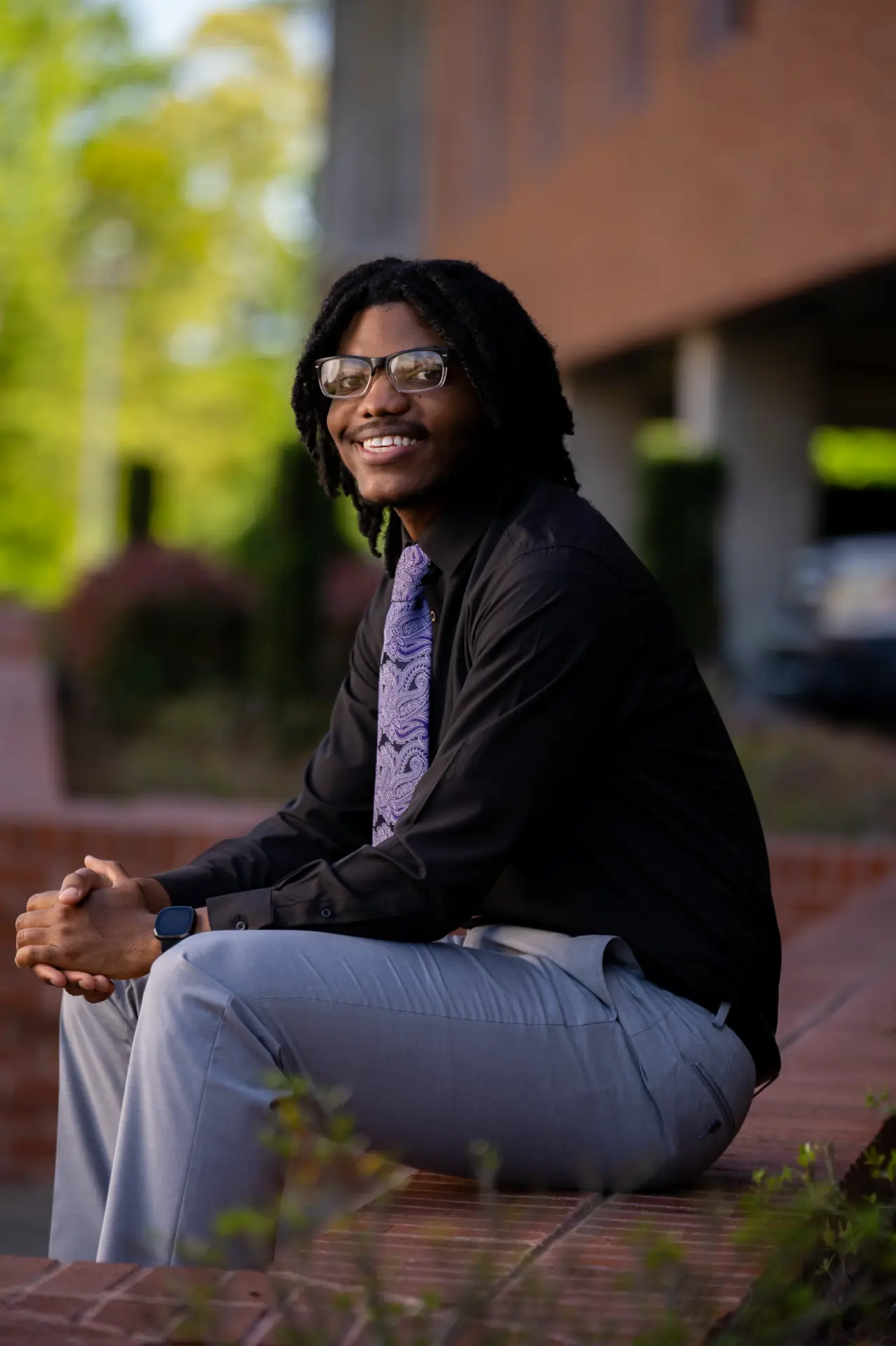 Graduate in black seated on a curb beside a campus building