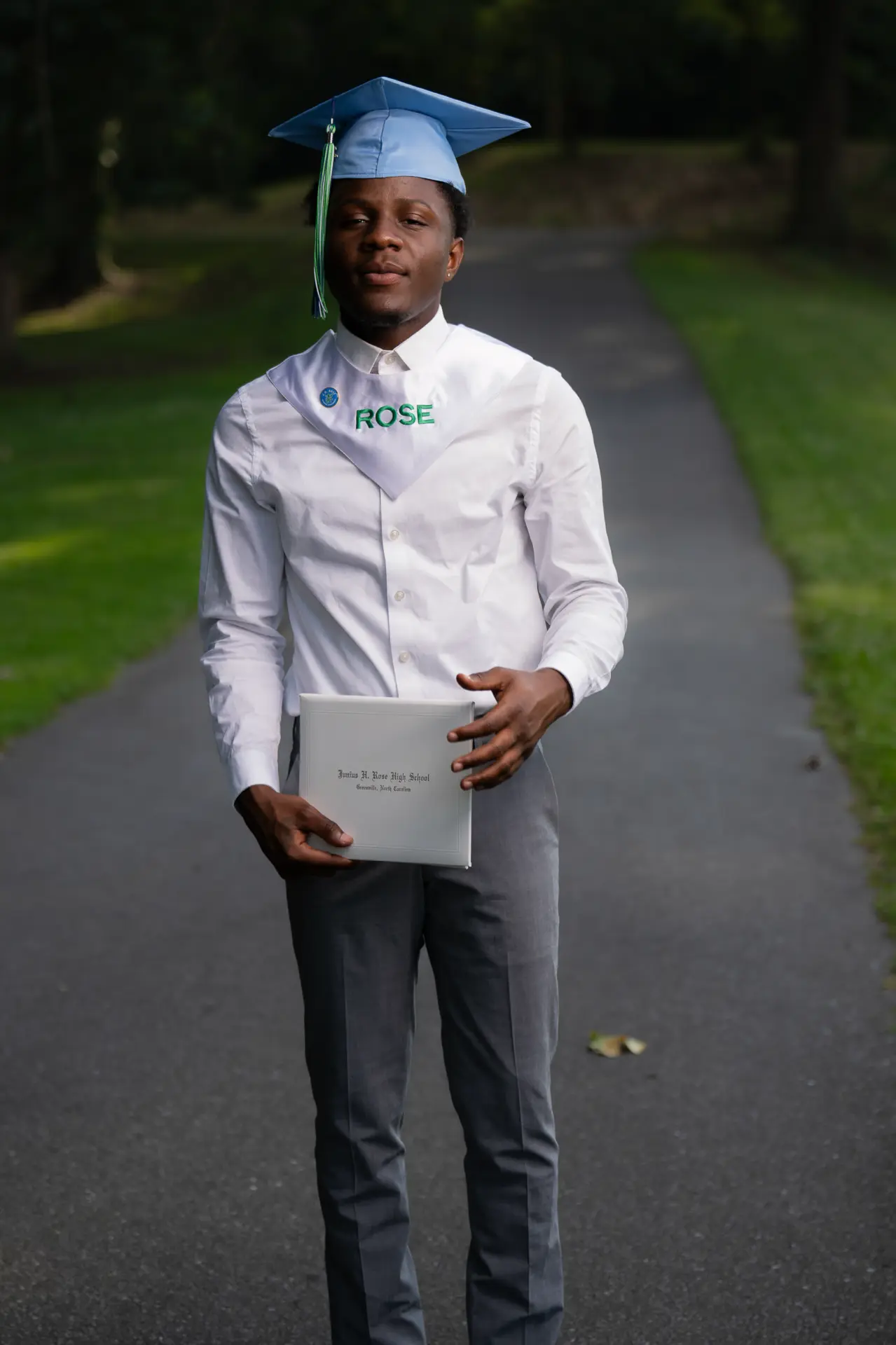 Male graduate in a white shirt and cap holding his diploma on a campus path