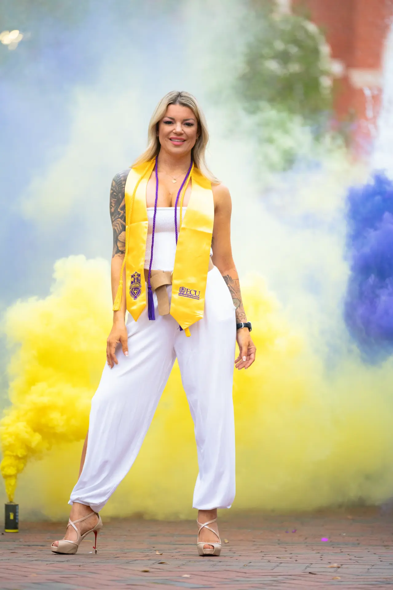 Graduate in white and gold stole standing in a cloud of yellow and purple smoke