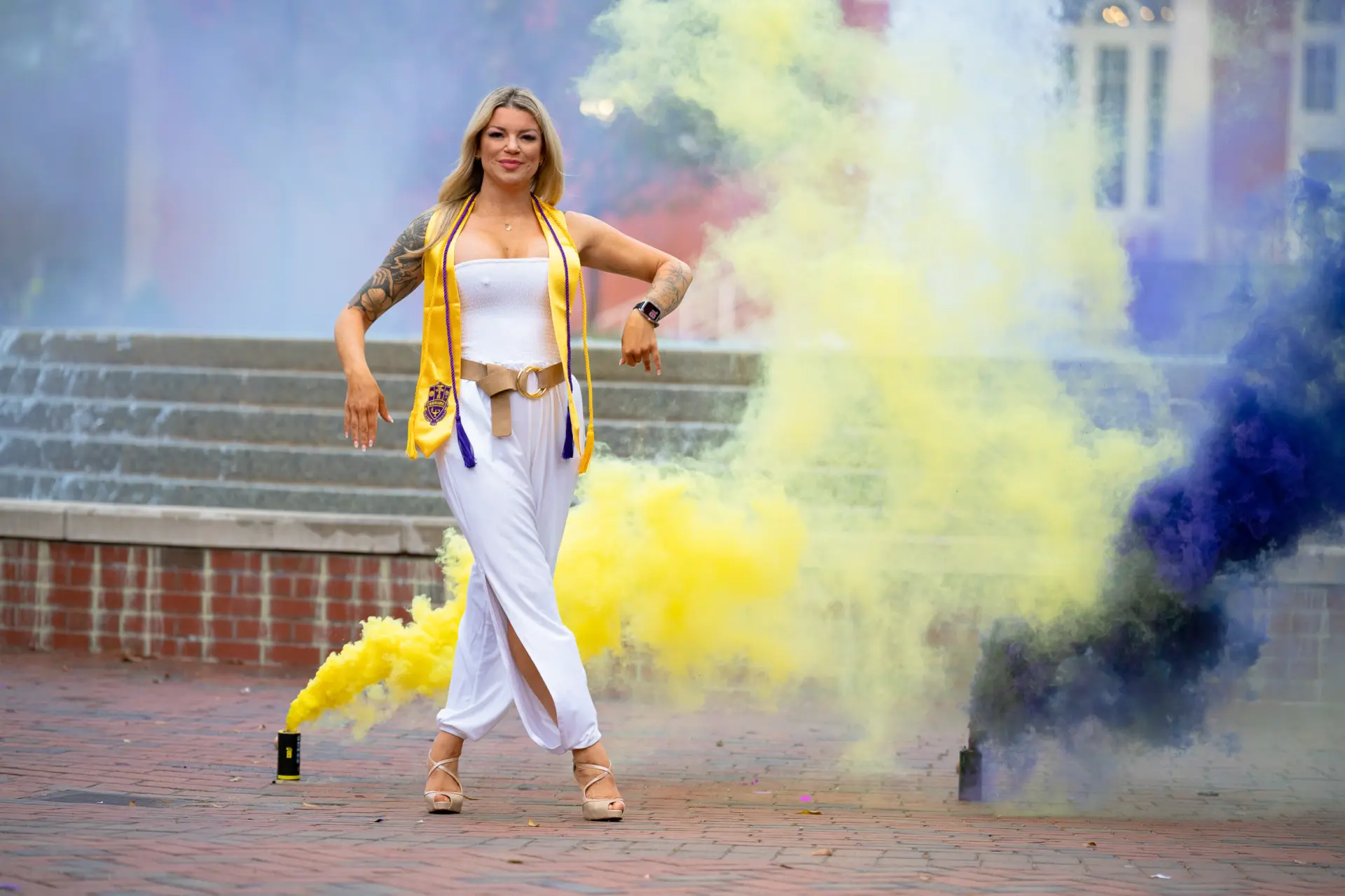 Graduate in white surrounded by purple and gold smoke bombs on campus