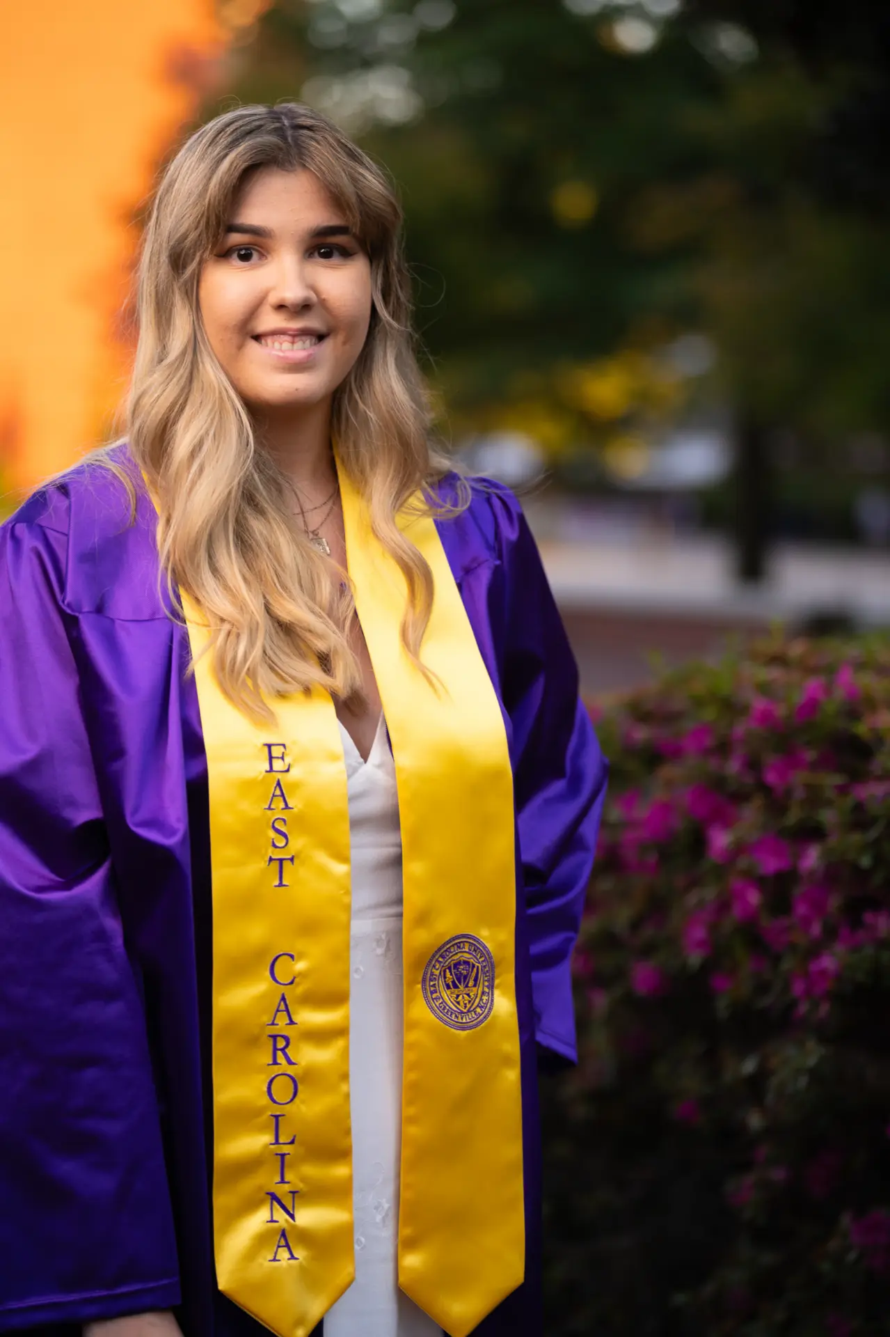 Graduate in a purple gown and gold stole by blooming campus shrubs