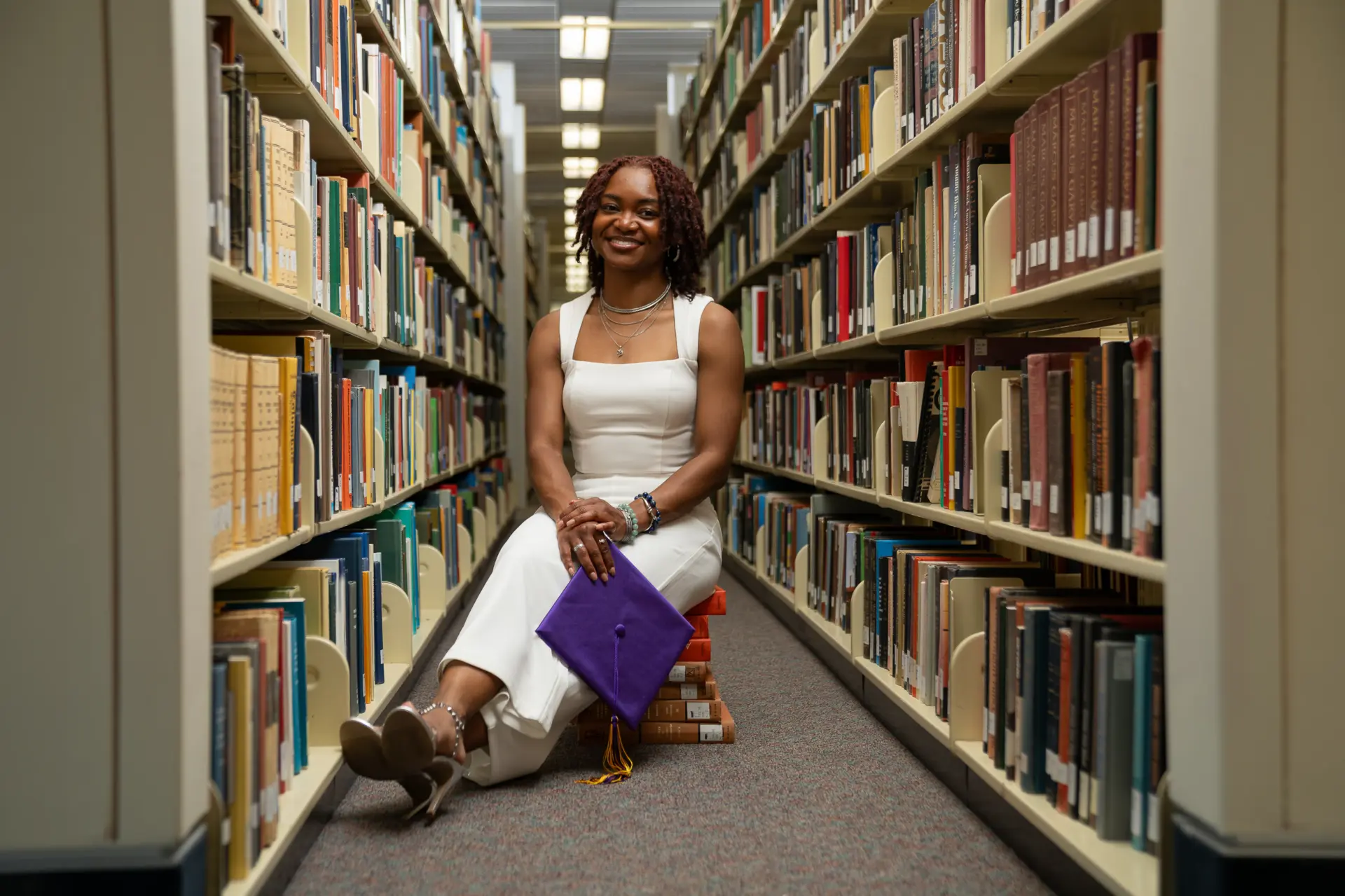 Graduate reading in a long library aisle between tall bookshelves