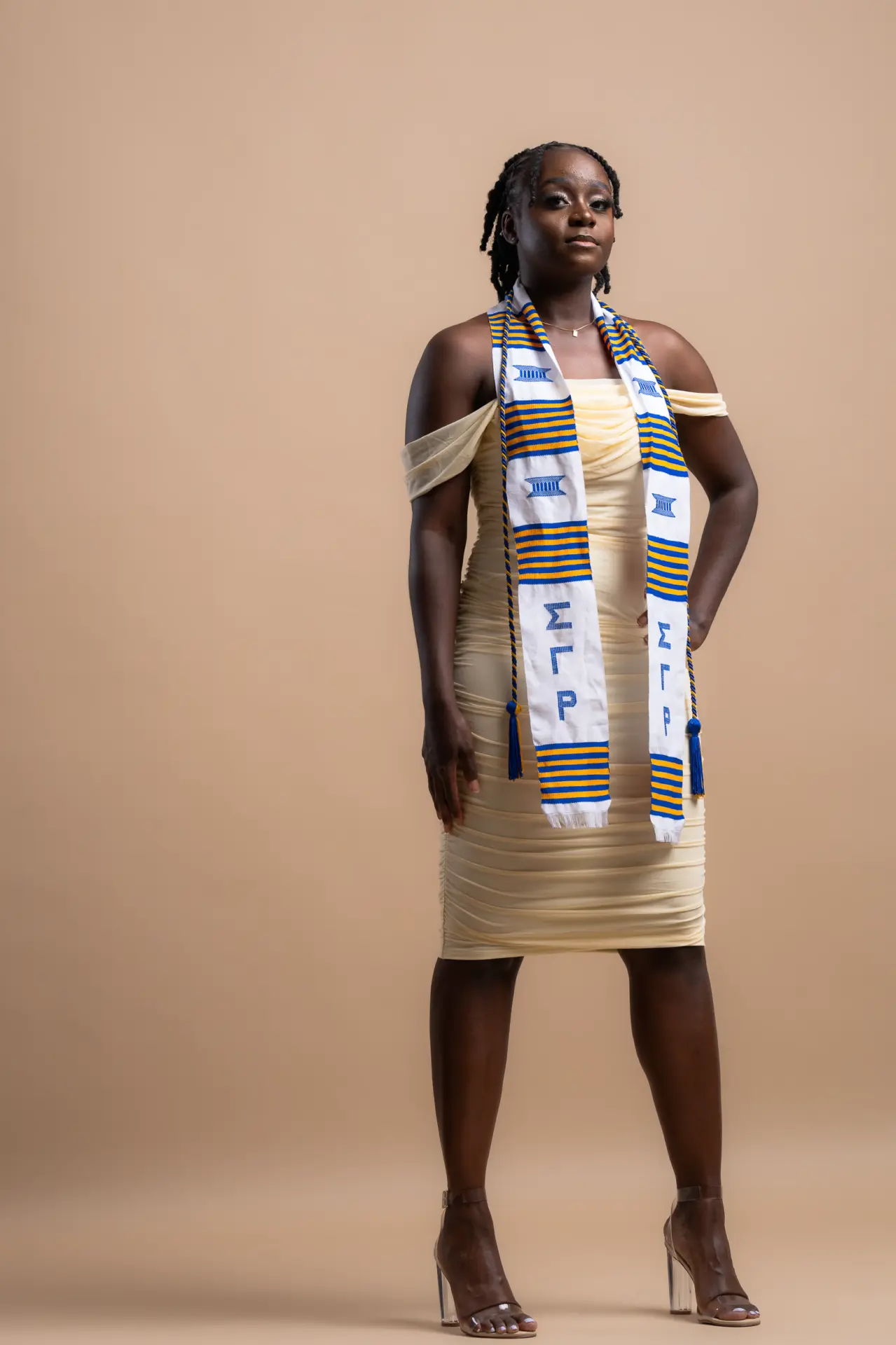 Graduate in a cream dress and stoles posing against a tan backdrop