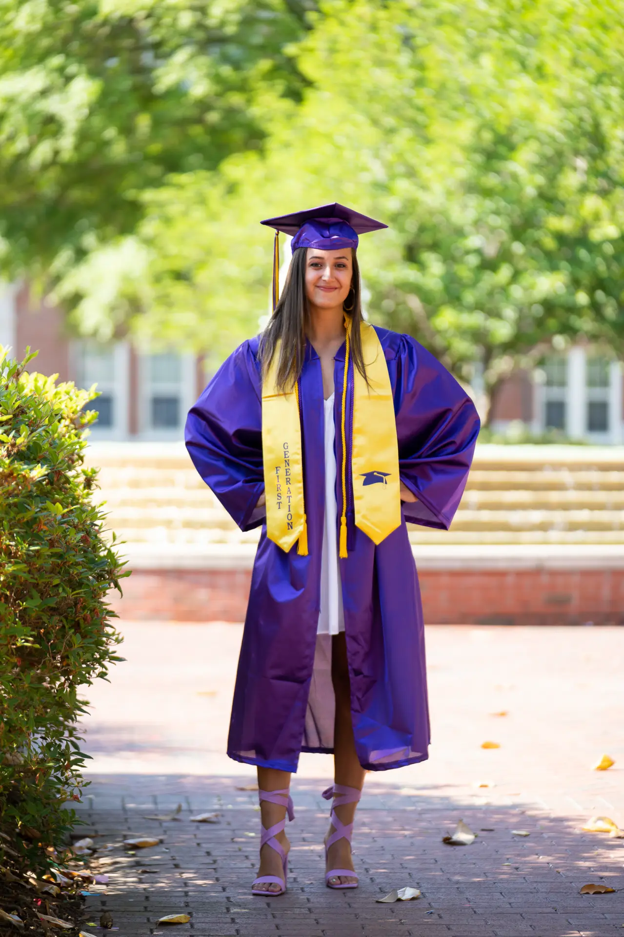 Graduate in purple cap and gown on a sunny brick walkway