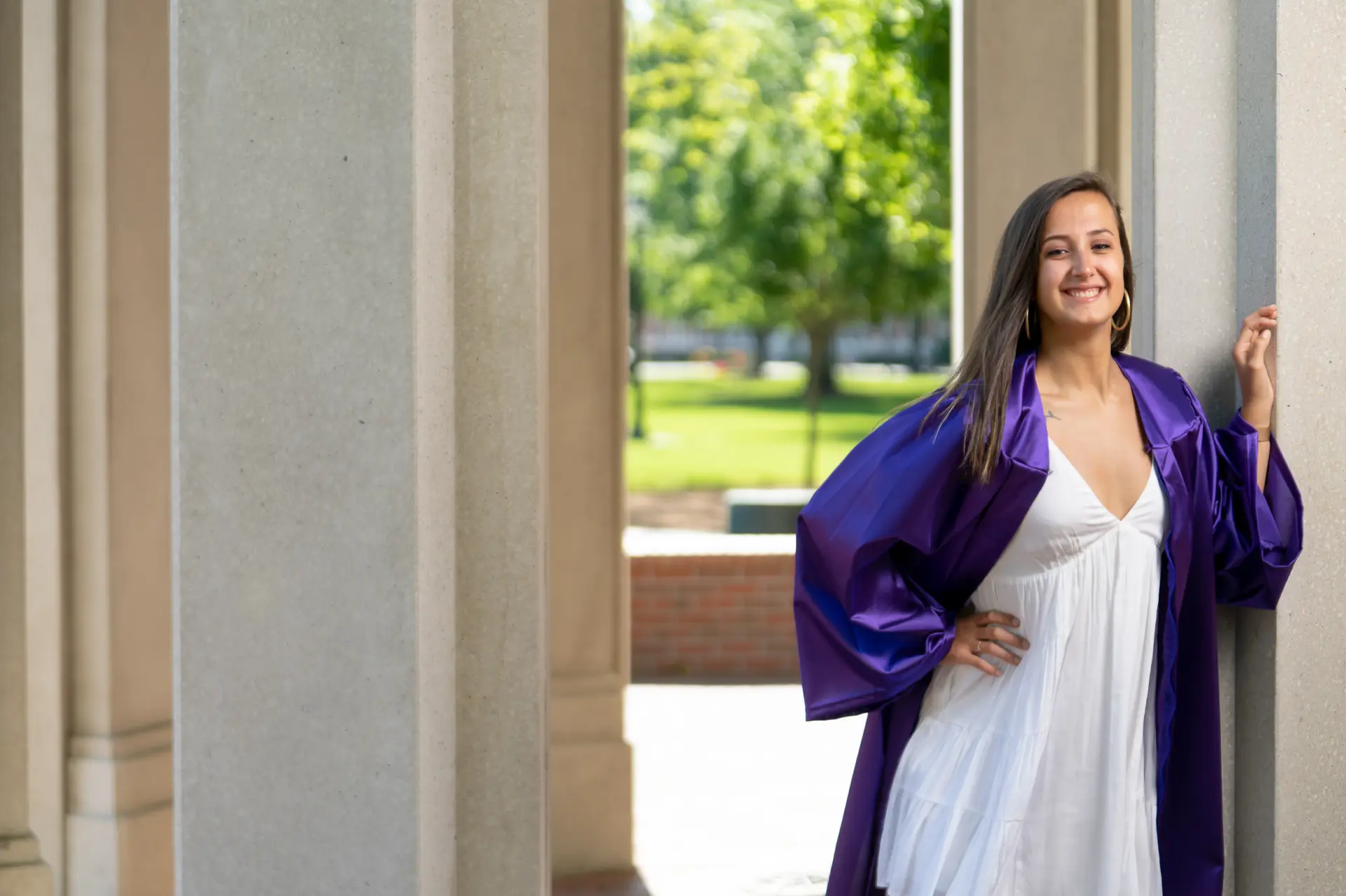 Graduate in a purple gown and white dress beside campus columns