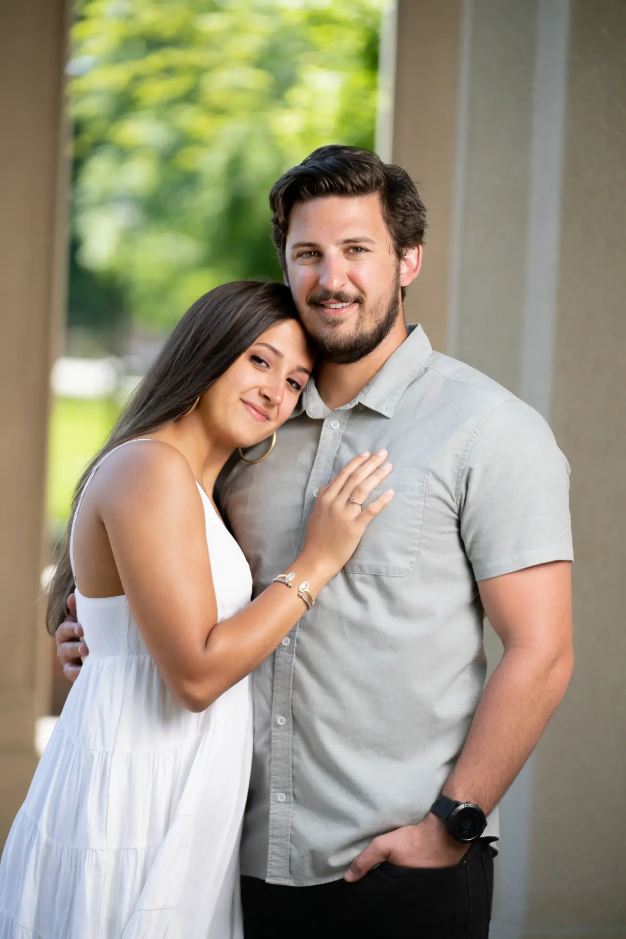 Graduate in white leaning on her partner's shoulder under the trees
