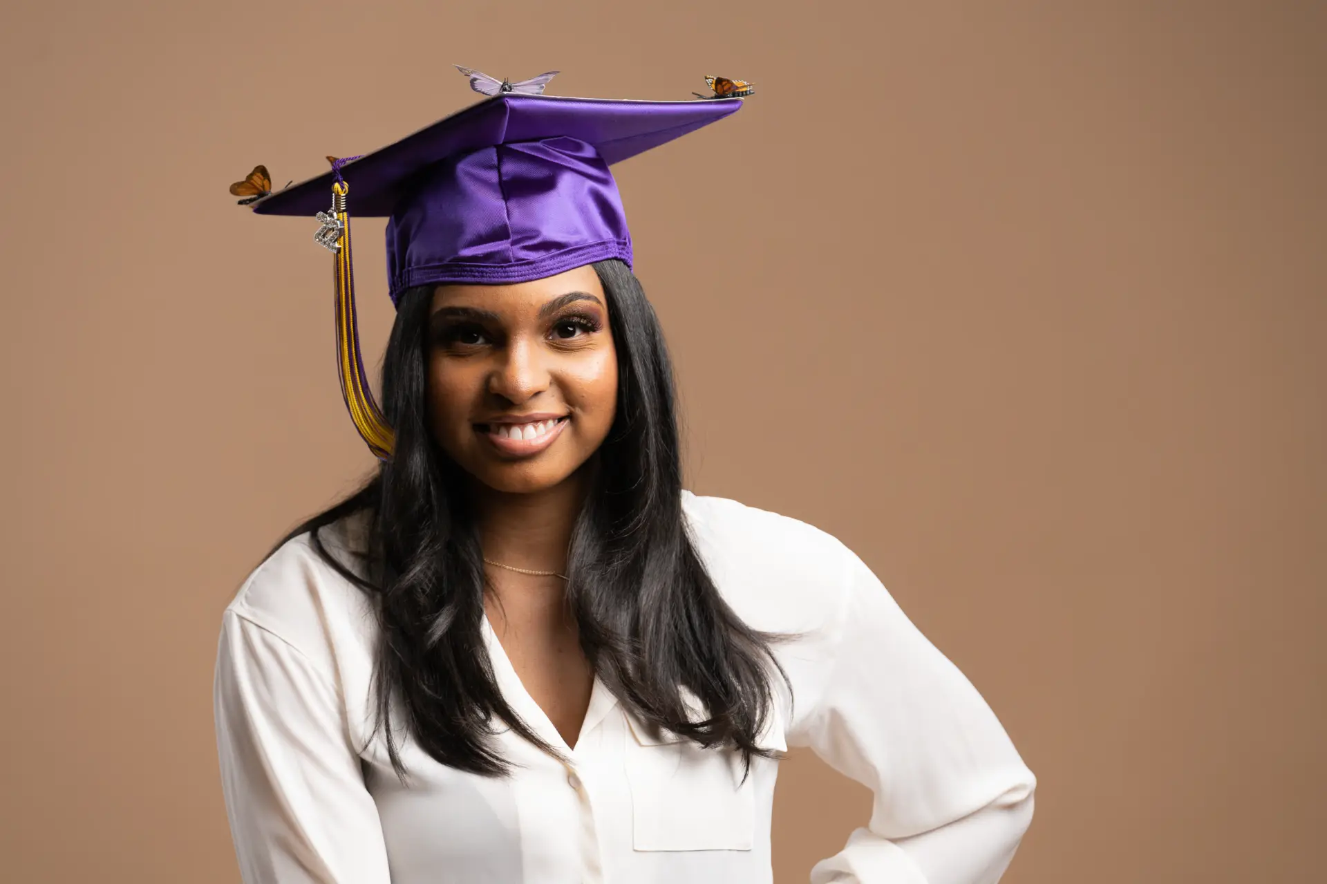 Smiling graduate in a purple cap against a warm tan backdrop