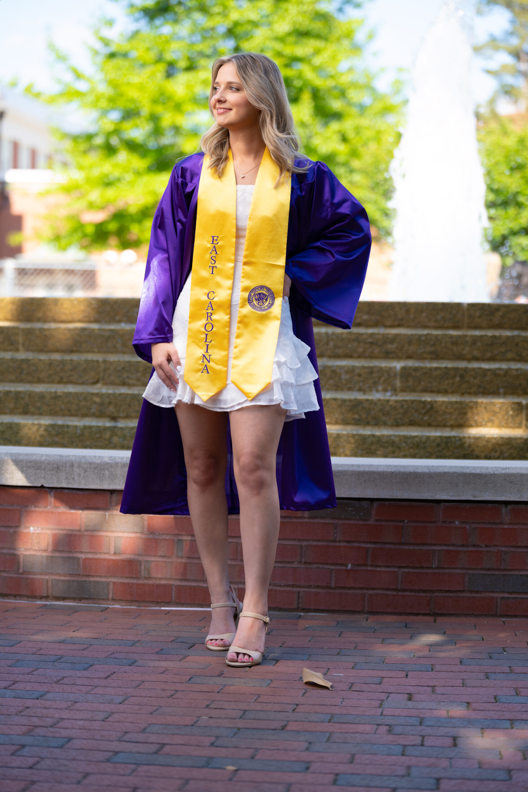 Graduate in a purple and gold stole posing by a campus fountain