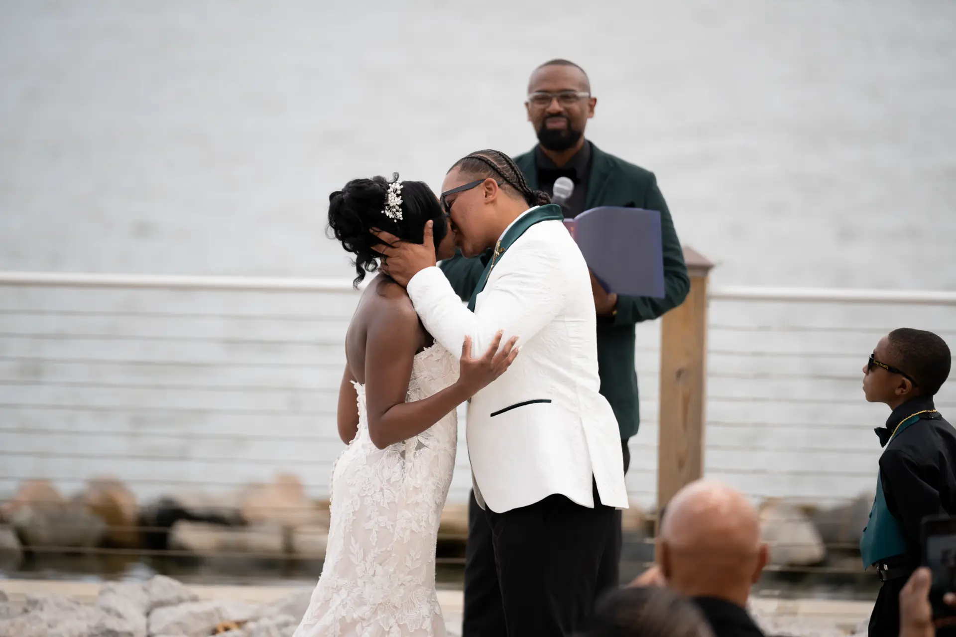 Groom in a white jacket embraces the bride by the water