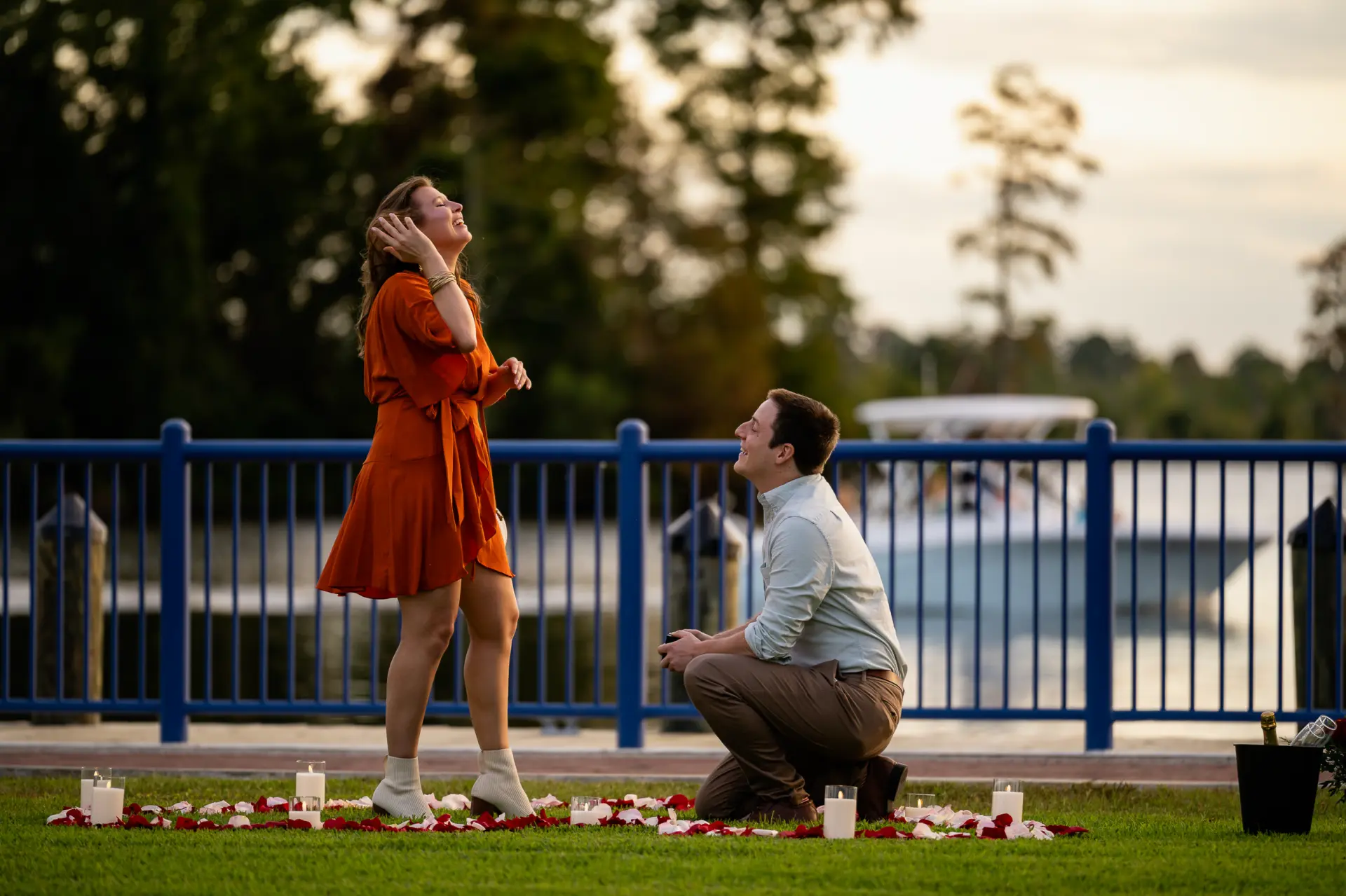 Man kneels to propose on a lakeside lawn lined with flowers