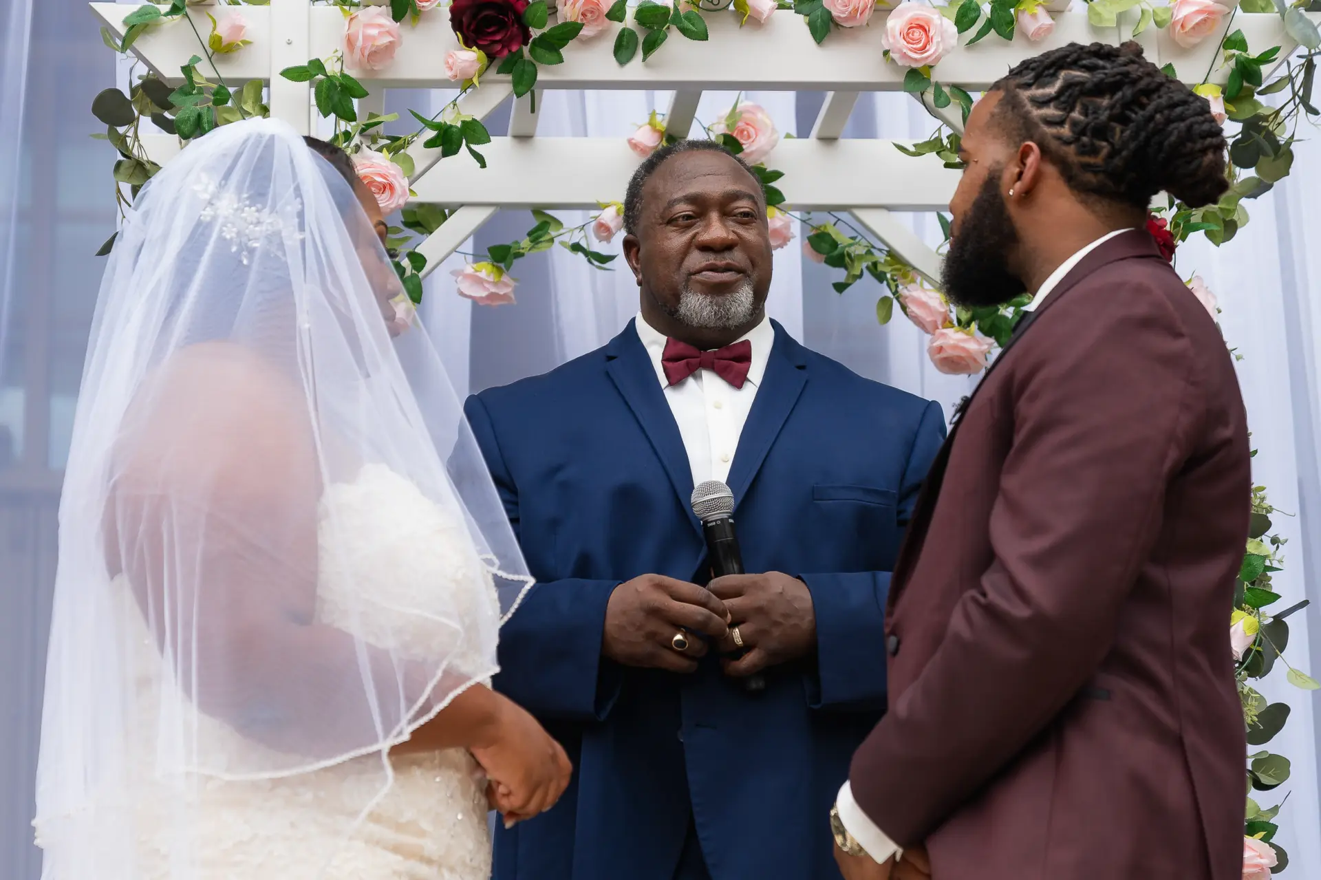 Bride in a veil smiles at the groom in a burgundy suit at the altar
